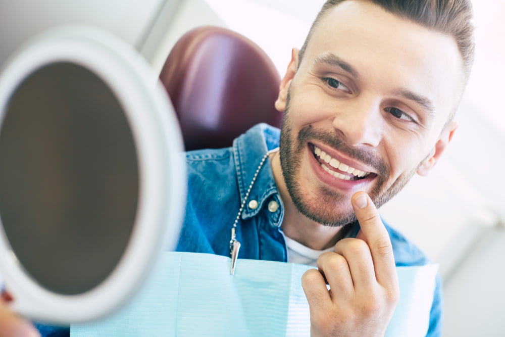 A man smiles and points to his teeth while looking at his reflection in a hand mirror during a dental visit - Dentist St George Utah A man smiles and points to his teeth while looking at his reflection in a hand mirror during a dental visit - Dentist St George Utah